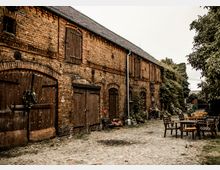Backsteinbau mit hölzernen Türen und Fenstern auf einem Kopfsteinpflasterhof, umgeben von Pflanzen und Blumenarrangements. Rechts im Bild stehen Gartenstühle und -tische, die eine rustikale Atmosphäre vermitteln.