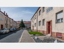 Wohnsiedlung mit zweistöckigen Reihenhäusern aus verputztem Mauerwerk und roten Ziegeldächern. Eine gepflasterte Straße führt durch die Wohnanlage, auf beiden Seiten sind geparkte Autos, Vorgärten mit Kiesflächen und ein zentraler Baum im Hintergrund sichtbar.