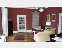 Wohnzimmer mit roten Wänden, einem Parkettboden und klassischen Möbeln. Ein Kachelofen befindet sich an der Wand, neben einem beigefarbenen Sofa, und der Raum ist mit Teppichen und Wanddekorationen geschmückt.