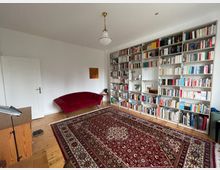 Wohnzimmer mit hellen Wänden, einer großen Wand mit Bücherregalen, die mit Büchern und Dekorationsgegenständen gefüllt sind. Auf dem Holzboden liegt ein großer, gemusterter Teppich, daneben ein rotes Sofa und eine einfache Deckenleuchte.
