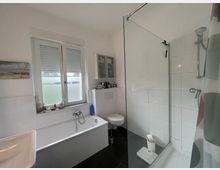 Modernes Badezimmer mit Badewanne, Glasduschkabine, Waschbecken und Toilette. Weiß geflieste Wände, dunkler Boden, großes Fenster mit Rollos und ein Hängeschrank an der Wand.