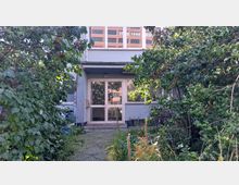 Eingangsbereich eines Wohngebäudes mit der Hausnummer 10, umgeben von dichter Vegetation und wild wachsenden Pflanzen. Im Hintergrund spiegeln sich moderne Gebäude in den Glasfenstern der Eingangstür.