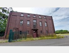 Backsteinbau mit rechteckiger Fassade, mehreren symmetrisch angeordneten Fenstern und einer zentralen, dunkelroten Eingangstür. Im Vordergrund ein grünes Metalltor, umgeben von Grasflächen und einer Straße, mit Bäumen und freiem Himmel im Hintergrund.