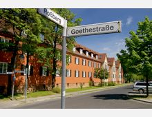 Straßenszene in einem Wohnviertel mit orangefarbenen Mehrfamilienhäusern, rotem Ziegeldach und weißen Fensterrahmen, umgeben von Bäumen. Im Vordergrund stehen Straßenschilder der "Goethestraße" und "Roßberg", daneben eine asphaltierte Straße und parkende Autos.