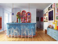 Bright, modern home bar area with a speckled blue counter, clear acrylic bar stools, and hanging pendant lights. Colorful arched wall shelves display bottles behind the bar, with light blue walls, wooden floors, and a gallery wall of framed artwork above a low cabinet.