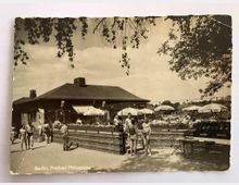 Historische Fotografie eines Freibads am Plötzensee in Berlin. Im Vordergrund sind Menschen in Badebekleidung, ein Gebäude mit Ziegelfassade und Schirmen im Außenbereich zu sehen, umgeben von Bäumen und weiteren Sitzgelegenheiten.