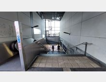Treppenaufgang mit angrenzender Rolltreppe in einem modernen, überdachten U-Bahn- oder Bahnhofsbereich. Die Wände sind mit grauen Platten verkleidet, und am Ende des Gangs befindet sich eine große Glasfront mit Blick nach draußen.