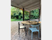 Überdachte Terrasse mit einer weißen Holzdecke, darunter ein runder Holztisch mit unterschiedlichen Stühlen auf einem gepflasterten Boden. Im Hintergrund ein Garten mit grünen Büschen, Bäumen und einer kleinen Rasenfläche, dekoriert mit bunten Lichterketten entlang der Überdachung.