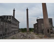 Alte Industrieanlage mit mehreren verfallenen Gebäuden, hohen Schornsteinen und Schutthaufen im Vordergrund, umgeben von vereinzelter Vegetation und bedecktem Himmel.
