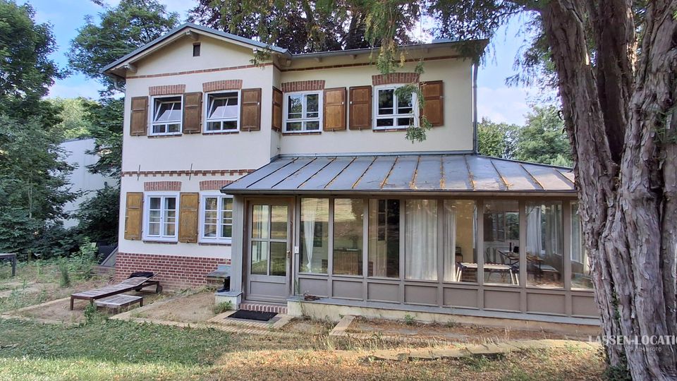 Zweistöckiges Wohnhaus mit hellem Putz, braunem Ziegelsockel und Fensterläden, davor ein verglaster Wintergarten mit Metallpultdach. Umgeben von einem Garten mit Rasen, Sitzmöbeln und hohen Bäumen am Waldrand.