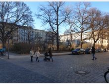 Straßenszene mit Kopfsteinpflaster, im Hintergrund mehrstöckige Wohnhäuser mit hellen Fassaden und dekorativer Architektur. Im Vordergrund stehen Bäume ohne Laub, parkende Autos und Personen, die spazieren gehen oder einen Kinderwagen schieben.