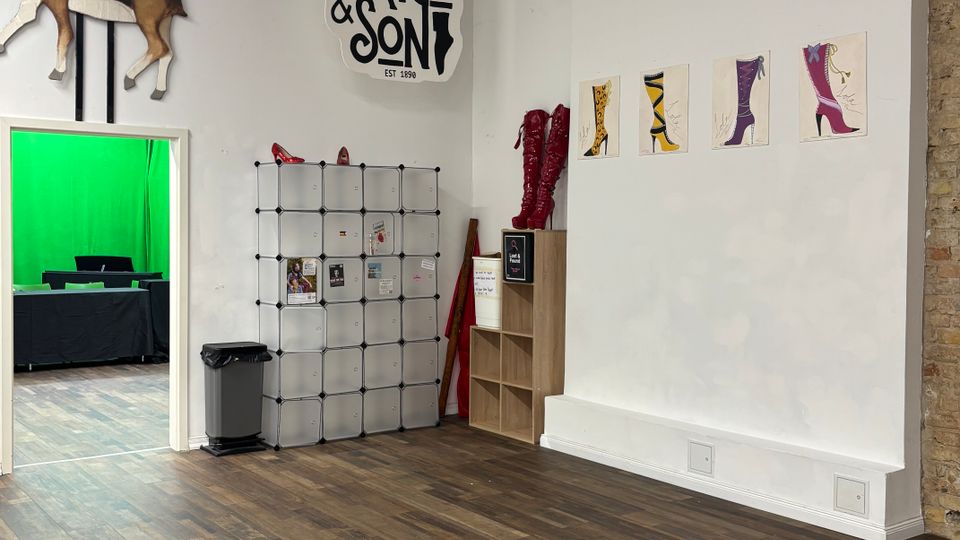 Interior of a studio or shop space with white walls, dark wood floors, and an exposed ceiling grid, featuring a “Price & Son” wall sign, a mounted antelope figure, and shoe-themed artwork. A doorway opens to a room with a green screen, and the wall area includes modular storage cubes and small shelving.