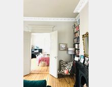 Wohnzimmer mit Holzdielenboden und Stuckdecke, geöffneten weißen Doppeltüren, die zu einem Esszimmer mit Klavier führen. Neben den Türen befindet sich ein Bücherregal und ein Stuhl mit Zebramuster; im Vordergrund ein Kamin mit Bilderrahmen und Dekorationen.