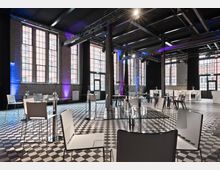 Großer, moderner Veranstaltungsraum mit hohen Decken, großen Industrie-Stil-Fenstern und schachbrettartigem Fliesenboden. Der Raum ist mit runden Tischen aus Glas, weißen Stühlen und dezent dekorierten Blumenvasen ausgestattet, wobei farbige Lichteffekte die Wände betonen.