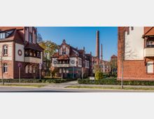 Straßenszene mit historischen Backsteingebäuden im späthistoristischen Stil, darunter Wohnhäuser mit dekorativen Fassadenelementen und Erkern. Im Hintergrund steht ein markanter Schornstein neben einer alten Industrieanlage.