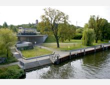 Parkähnliche Anlage mit einer Uferpromenade an einem Fluss, einem großen Baum und einer Weide. Im Hintergrund eine Brücke mit einem "Potsdam City Tour"-Doppeldeckerbus und ein Turm auf einem Hügel.