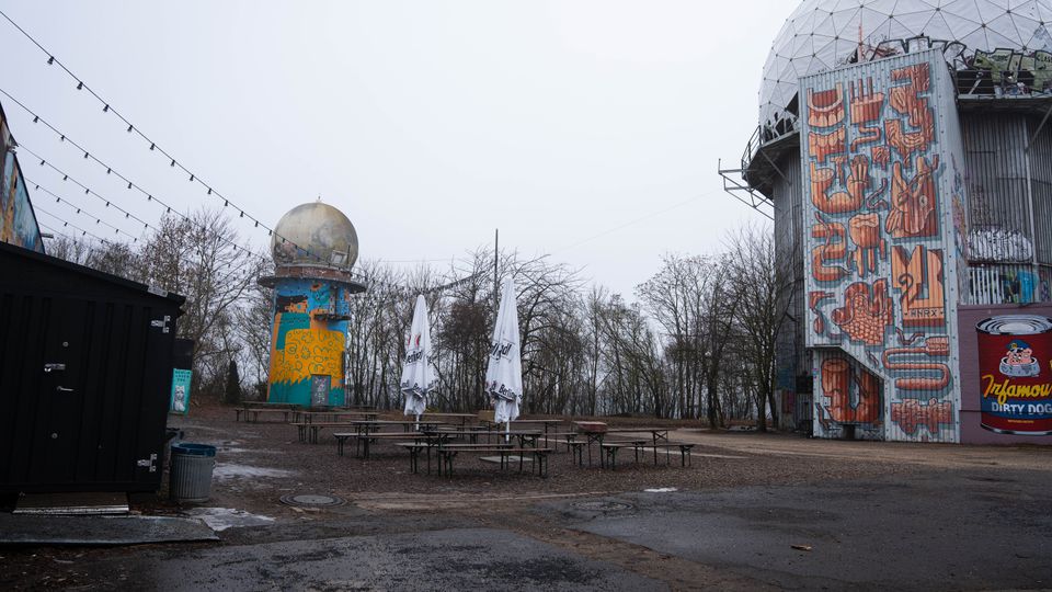 Offener Außenbereich mit Bierbänken und geschlossenen Sonnenschirmen auf einem unbefestigten Platz, umgeben von kahlen Bäumen. Rechts steht ein hoher, zylindrischer Bau mit großer geodätischer Kuppel und Graffiti-Fassade, links ein kleinerer Turm mit Kugelaufsatz und bunter Bemalung; über dem Platz hängen Lichterketten.