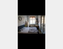 Kleines Schlafzimmer mit einem Doppelbett, hellem Holzboden und einem großen Fenster, das viel Tageslicht hineinlässt. Im Raum befinden sich außerdem ein Schreibtisch, ein Regal mit Ordnern sowie ein Wandspiegel.