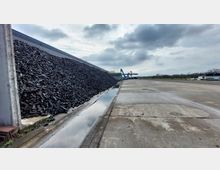 Ein Industriegelände mit einem großen Haufen schwarzer Kohle links, einer Betondecke mit Regenwasserpfützen und blauen Industriekränen im Hintergrund. Der Himmel ist bewölkt, und die Umgebung wirkt offen mit vereinzelten Bäumen in der Ferne.