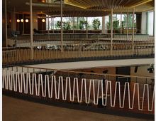 Innenansicht eines modernen Gebäudes mit mehreren Etagen, die durch offene Galerien verbunden sind. Charakteristisch sind die dekorativen Geländer mit weißen, geometrischen Elementen und große Fenster mit Blick auf grüne Pflanzen.