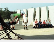 Ein Skatepark mit Jugendlichen, von denen einer auf Inlineskates eine Rampe herabfährt. Im Hintergrund sind moderne Betonwände und Bäume zu sehen, während andere Jugendliche auf einer Bank sitzen und zuschauen.