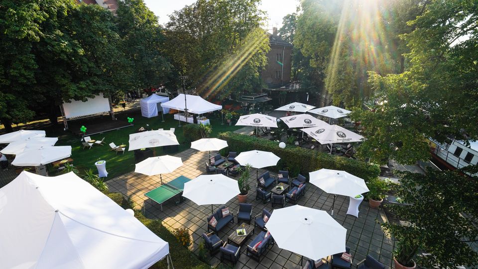 Luftaufnahme eines offenen Biergartens oder Veranstaltungsortes mit weißen Sonnenschirmen, Sitzgelegenheiten auf einer gepflasterten Fläche und einem großen grünen Rasenbereich. Im Hintergrund stehen große Bäume, Zelte und ein Boot, während Sonnenstrahlen durch die Baumkronen scheinen.