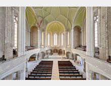 Innenraum einer historischen Kirche mit hohen Rundbogenfenstern, einem grünen Gewölbedeckenbereich und sichtbarer Alterung der Wände. In der Mitte befindet sich ein Altar mit einem großen Holzkreuz, umgeben von Holzbänken und einer umlaufenden Empore.