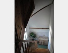 Treppenhaus mit Holztreppe und gedrechseltem Geländer unter einer hohen, schrägen Decke mit sichtbaren Holzbalken. Neben einem Fenster stehen eine Topfpflanze und eine niedrige Holzkommode auf grünem Teppichboden.