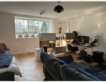 Modern eingerichtetes Wohnzimmer mit einem großen Fenster, das Tageslicht hereinlässt. Der Raum verfügt über einen Schreibtisch mit Computer, ein offenes Regal mit Figuren, ein graues Sofa, ein Bett und Holzparkettboden.