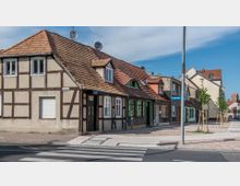 Fachwerkhäuser mit roten Ziegeldächern an einer Straßenecke, markiert durch ein Straßenschild "Feldstraße". Im Vordergrund sind ein Zebrastreifen und ein gepflasterter Gehweg mit neu gepflanzten Bäumen zu sehen.