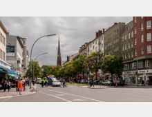 Eine städtische Straße mit mehrstöckigen Gebäuden, Geschäften und Wohnhäusern auf beiden Seiten; im Hintergrund ist die Spitze eines Kirchturms zu sehen. Auf der Straße stehen Passanten, Radfahrer sowie ein Krankenwagen, während links ein kleiner Marktbereich zu erkennen ist.