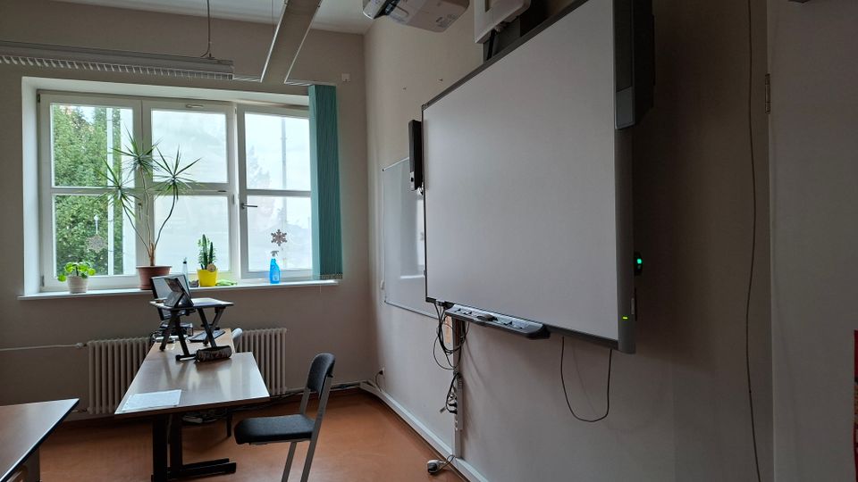 Klassenzimmer mit großem Fenster, mehreren Topfpflanzen auf der Fensterbank und einem Lehrerpult mit Laptop. An der Wand hängt ein großes interaktives Whiteboard mit Kabeln und darüber ein Deckenprojektor.