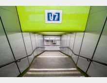 Treppenabgang zu einer U-Bahn-Station der Linie U7, mit hellgrünen Wänden und Handläufen an beiden Seiten. Im Hintergrund sind der Bahnsteigbereich und Informationsschilder zu sehen.