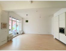 Heller, leerstehender Raum mit weiß gestrichenen Wänden, großen Fenstern auf der linken Seite, hellem Holzfußboden und einem eingebauten Küchenschrank mit Backofen auf der rechten Seite. An der Decke hängen zwei minimalistische Lampen, und durch die Fenster sind Balkone und umliegende Gebäude sichtbar.