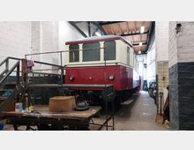 Roter und cremefarbener Zugwagon in einer Werkstatthalle mit weißen Wänden und offener Holzbalkendecke. Im Vordergrund stehen Arbeitsgeräte und Materialien auf einem Tisch, während der Raum von Beleuchtung und Werkzeugen entlang der Wände geprägt ist.