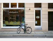 Außenansicht eines Geschäfts mit der Aufschrift "Schokogalerie" auf der verglasten Schaufensterfront, daneben eine weiße Eingangstür. Vor der Fassade steht ein schwarzes Fahrrad, das an einem Laternenpfahl angelehnt ist.