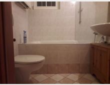 Badezimmer mit beigefarbenen Fliesen an Wand und Boden, einer Badewanne mit Duschbrause, einer Toilette und einem Holzunterschrank mit Waschbecken. Im oberen Bereich befindet sich ein kleines Fenster.