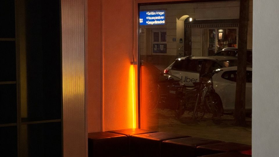Innenraum eines kleinen Eingangs- oder Wartebereichs mit gefliester Bodenfläche, gepolsterten Sitzbänken und warmem orangefarbenem Licht. Eine große Fensterfront zeigt den Blick auf die Straße mit geparkten Autos und Fahrrädern; darüber sind Lüftungselemente und eine Deckenleuchte zu sehen.