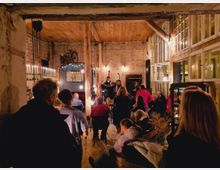 Ein rustikaler Raum mit Backsteinwänden und Holzbalkendecke, in dem ein Live-Auftritt stattfindet. Menschen sitzen und stehen, während ein Musiker vorne mit Mikrofon und Gitarre zu sehen ist; von der Decke hängen Fahrräder und Lampen als Dekoration.