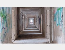Langer, verlassener Flur in einem heruntergekommenen Gebäude, dessen Wände mit Graffiti bedeckt sind; der Boden ist uneben, und am Ende des Flurs ist ein fensterartiger Lichtausschnitt sichtbar.