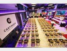 Veranstaltungsraum mit Reihen aus hölzernen Stühlen, einer Sitzbank mit Kissen an der Wand und einem großen Schild mit der Aufschrift "Cantinerie". Im Hintergrund befinden sich gedeckte Tische, eine Bar und dekorative Elemente wie Blumenarrangements.