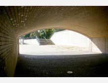 Blick durch eine Bogenunterführung aus gelben Klinkersteinen mit gepflastertem Boden, die zu einem hellen, offenen Bereich mit Treppen und einem Geländer führt. Im Hintergrund sind Bäume sichtbar.
