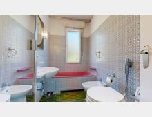 Kleines Badezimmer mit rosa Badewanne, weißem Waschbecken, Toilette und Bidet. Die Wände sind mit gemusterten Fliesen verkleidet, der Boden hat grüne Keramikfliesen, und ein Fenster mit Milchglas lässt Tageslicht herein.