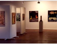 Innenraum einer Kunstgalerie mit weißen Trennwänden, an denen Gemälde hängen. Im Hintergrund stehen ein Podest mit einer kleinen Skulptur und weitere Kunstwerke an der Wand. Die Beleuchtung ist warm und fokussiert auf die Ausstellungsstücke.