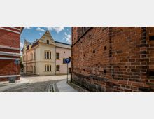 Pflastersteinstraße in einer historischen Altstadt mit einem gelb-beigen Gebäude im Stil der Renaissance und roten Backsteinfassaden auf beiden Seiten. Ein Verkehrsschild mit einem blauen Pfeil nach oben steht am Straßenrand, und die Szene ist bei Tageslicht aufgenommen.