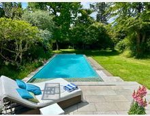 Ein privater Garten mit einem rechteckigen Pool, umgeben von grüner Rasenfläche und üppigen Bäumen. Im Vordergrund steht eine Liege mit blauen Kissen und einem Handtuch auf einer gepflasterten Terrasse.