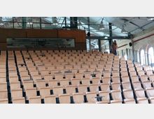 Ein moderner Hörsaal mit ansteigenden Holzsitzen, ausgestattet mit kleinen Klapptischen. Im Hintergrund sind Ziegelwände mit Rundbogenfenstern und einer Glasfront unter einem Metallträgerdach zu sehen.