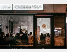 Außenansicht eines Veranstaltungsraums mit gläserner Eingangstür, hinter der mehrere Personen in einem minimalistischen Innenraum mit rauen Wänden sitzen; oben ein Schild mit Veranstaltungsinformationen.