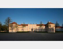 Helles, symmetrisches Schlossgebäude mit drei Flügeln und rotem Ziegeldach, umgeben von einer offenen Kiesfläche und Bäumen im Vordergrund. Der blaue Himmel und die tiefstehende Sonne erzeugen lange Schatten auf der cremefarbenen Fassade.