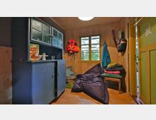 Holzvertäfelter Innenraum mit einem blauen Schrank auf der linken Seite, einem kleinen Fenster in der Mitte und einer gelben Eingangstür rechts. Im Raum befinden sich eine Sitzgelegenheit mit Kissen, ein großer Sitzsack und an der Wand aufgehängte Jacken sowie ein Gewehr.